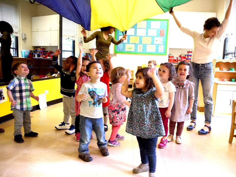 Several toddlers beneath a colorful canopy held up by students